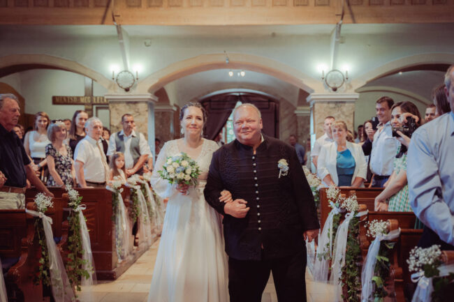 A menyasszony édesapjával vonul az oltár felé a zsúfolásig telt vidéki templomi esküvőn.
