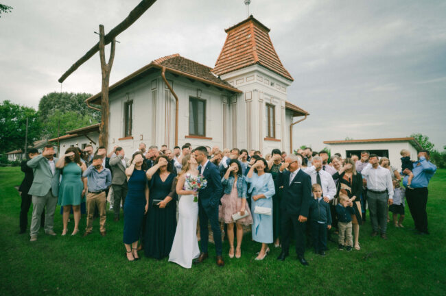 Vidám nagycsoportkép a teljes násznéppel egy szabadtéri esküvőn, történelmi épülettel a háttérben.