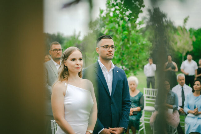 A menyasszony és a vőlegény egymás kezét fogva állnak a meghitt szabadtéri esküvői szertartáson.